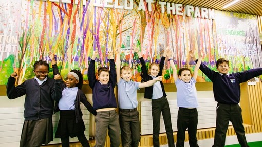 A group of children lined up in front of a large painting on the wall. They are smiling and throwing their arms up in the air. 