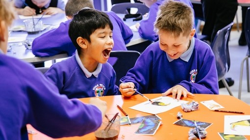 An image of two young people sat on their desk, painting. One child is laughing whilst the other has his head down, concentrating on his painting. 