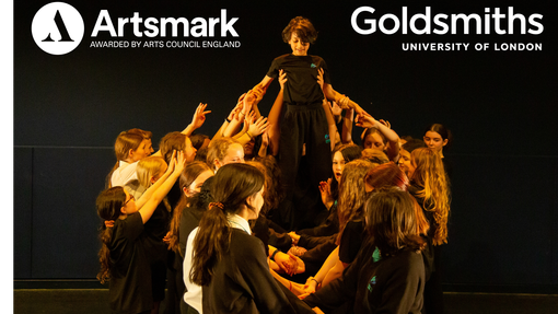 A group of students on a dark stage holding up another student, who is standing on their arms. Artsmark's logo is top left, Goldsmiths logo is top right